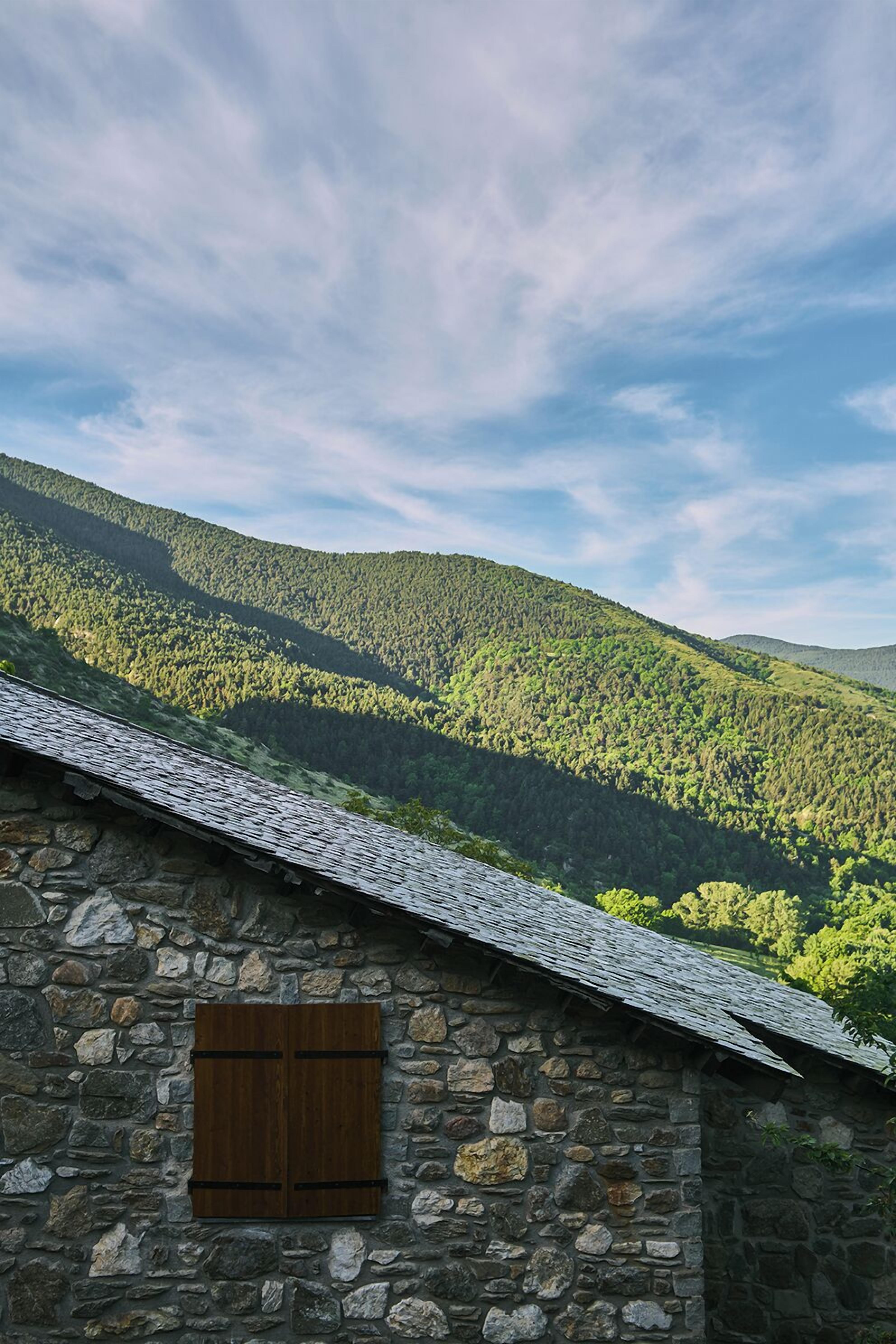 Un edificio de piedra con una persiana de madera, con un paisaje de colinas boscosas de fondo bajo un cielo parcialmente nublado.