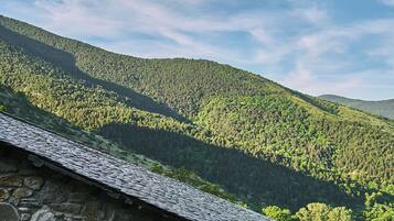 Un edificio de piedra con una persiana de madera, con un paisaje de colinas boscosas de fondo bajo un cielo parcialmente nublado.