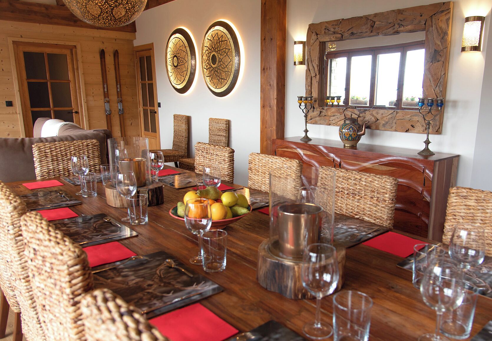 Un comedor con una mesa de madera dispuesta para seis personas, rodeada de sillas de mimbre, con un frutero y copas de vino sobre la mesa.