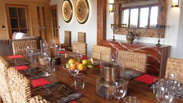 Un comedor con una mesa de madera dispuesta para seis personas, rodeada de sillas de mimbre, con un frutero y copas de vino sobre la mesa.