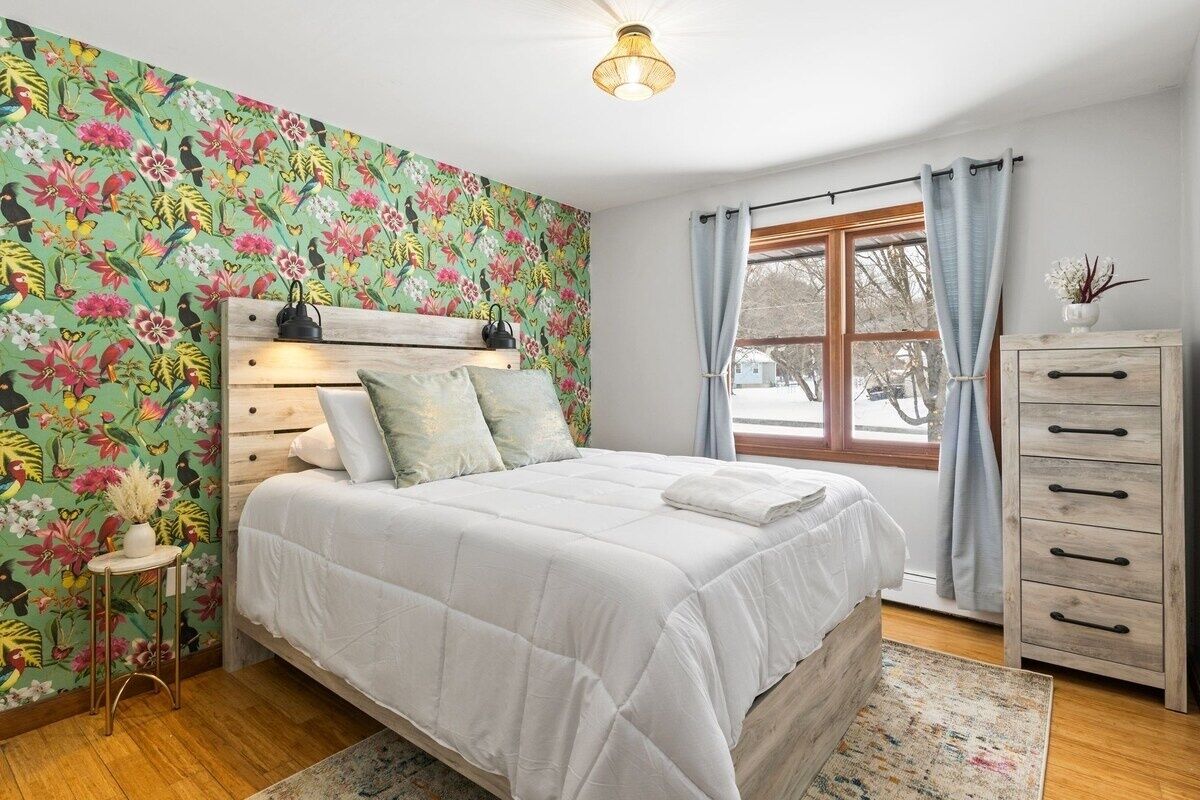 Un dormitorio con papel tapiz floral, un cabecero de madera, una mesita de noche, un aparador y una ventana con cortinas.