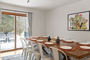 A dining room with a large table set for six, white chairs, and a sliding glass door leading to an outdoor area with chairs.
