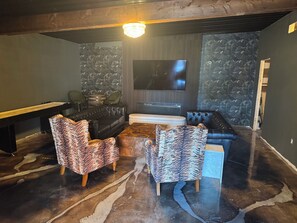 A modern living room with a black leather sofa, two patterned armchairs, a coffee table, and a flat-screen TV mounted on the wall.