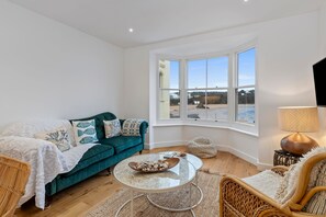 A modern living room with a teal sofa, a glass coffee table, wicker chairs, and a large window offering a view of the outdoors. - Azure - Sea Views Town Centre (Tenby)