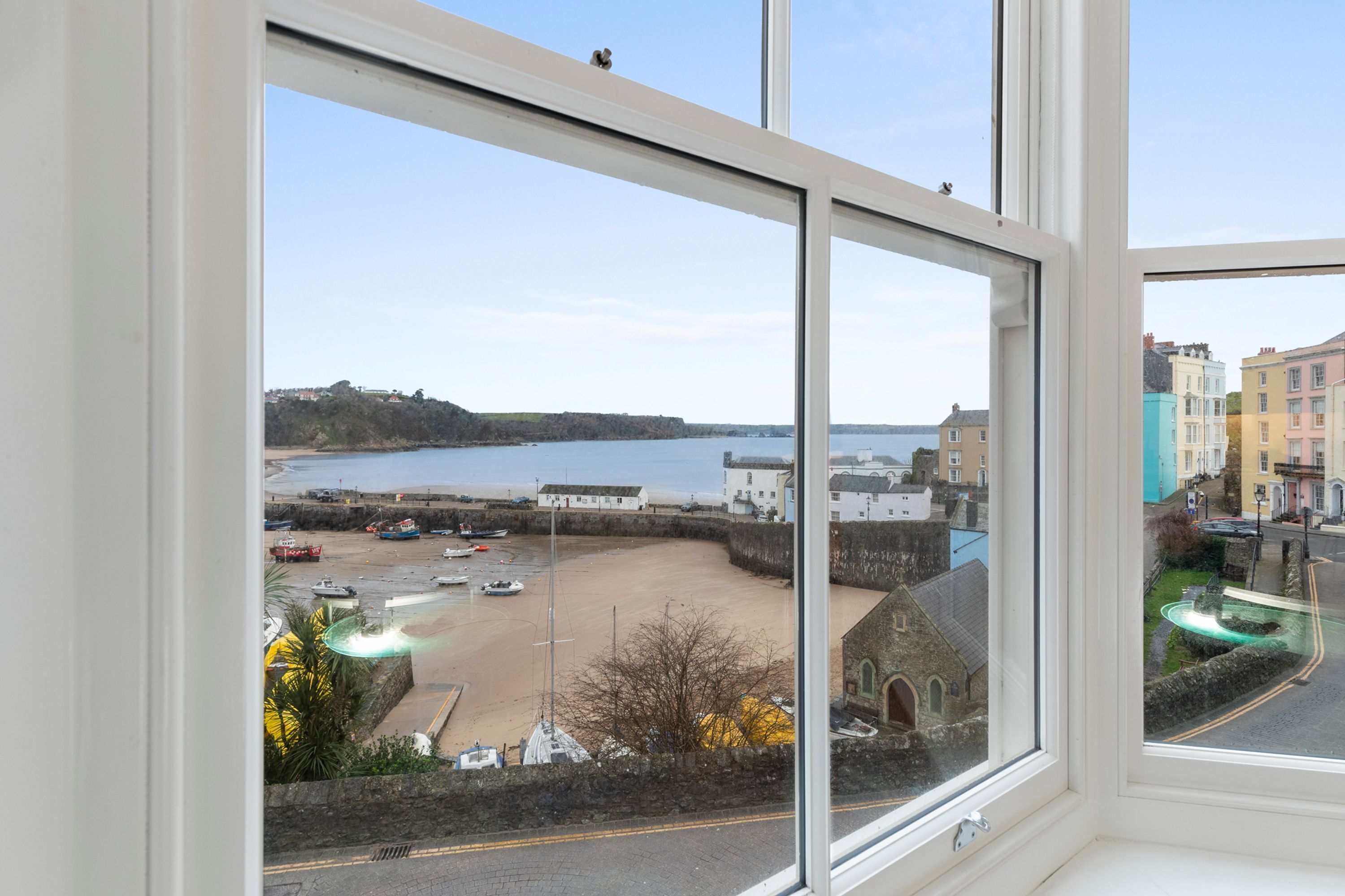 A coastal view from a window overlooking a sandy beach with boats, a stone wall, and buildings in the background.