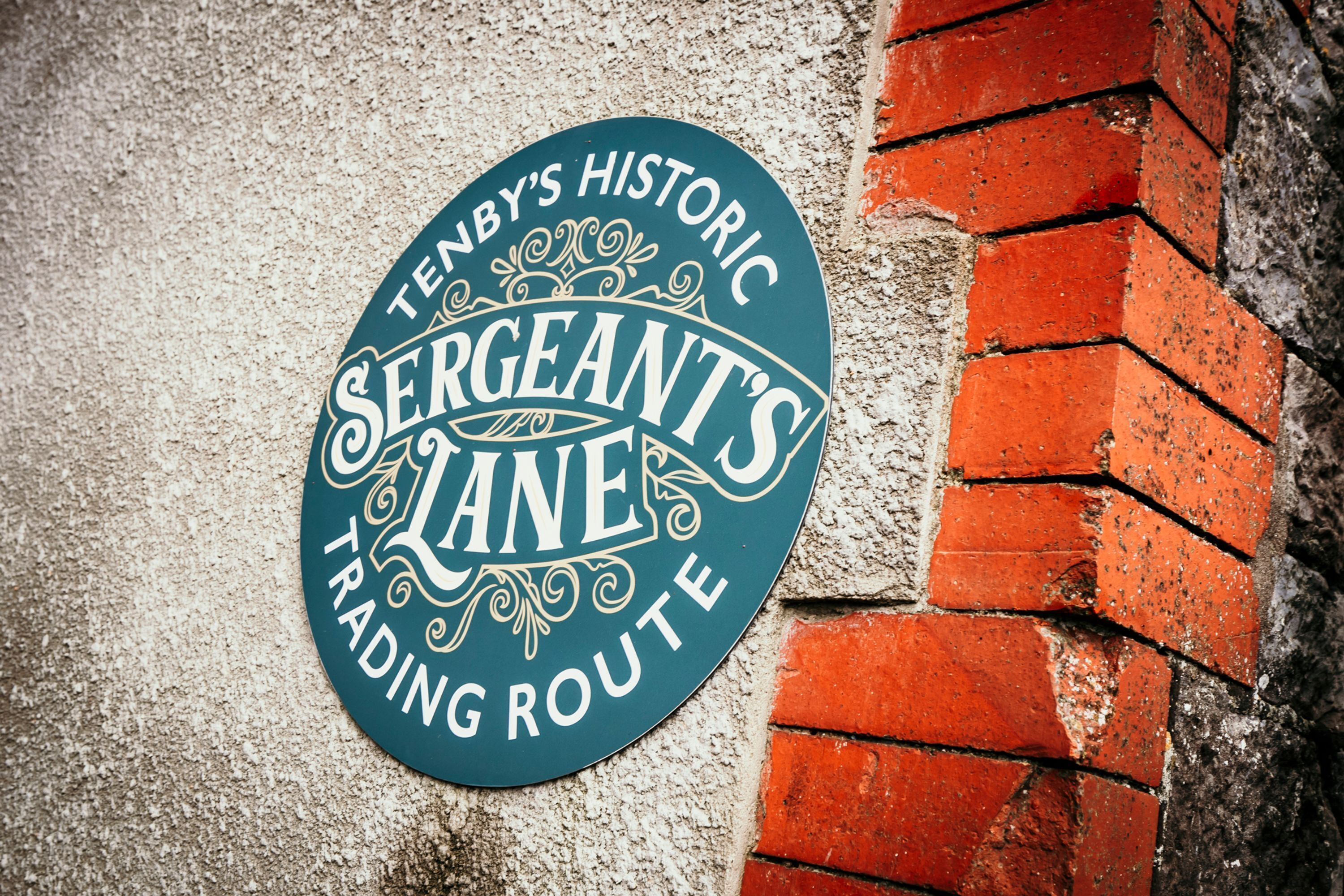 A circular sign with the text "Sergeant's Lane" and "Tenby's Historic Trading Route" against a brick wall.
