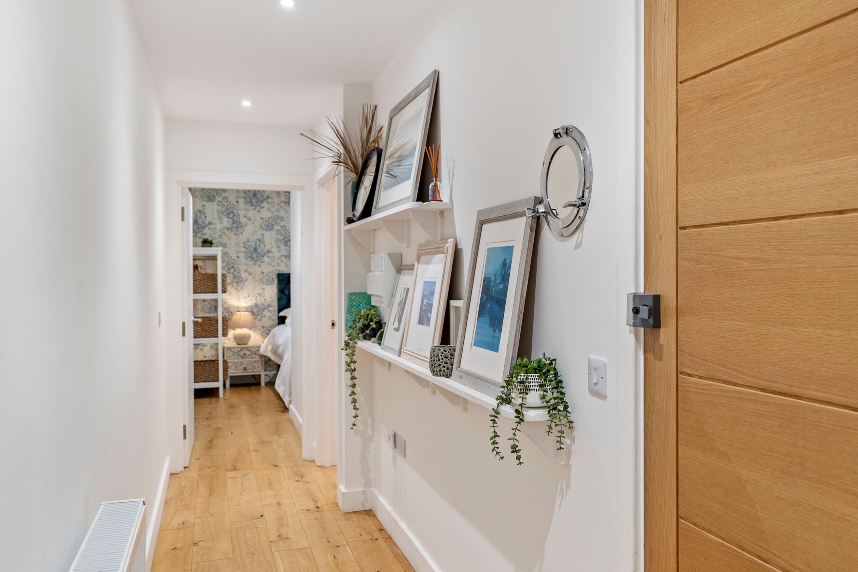 A hallway with a wooden door, a white wall with a shelf holding framed pictures and a mirror, and a wooden floor.