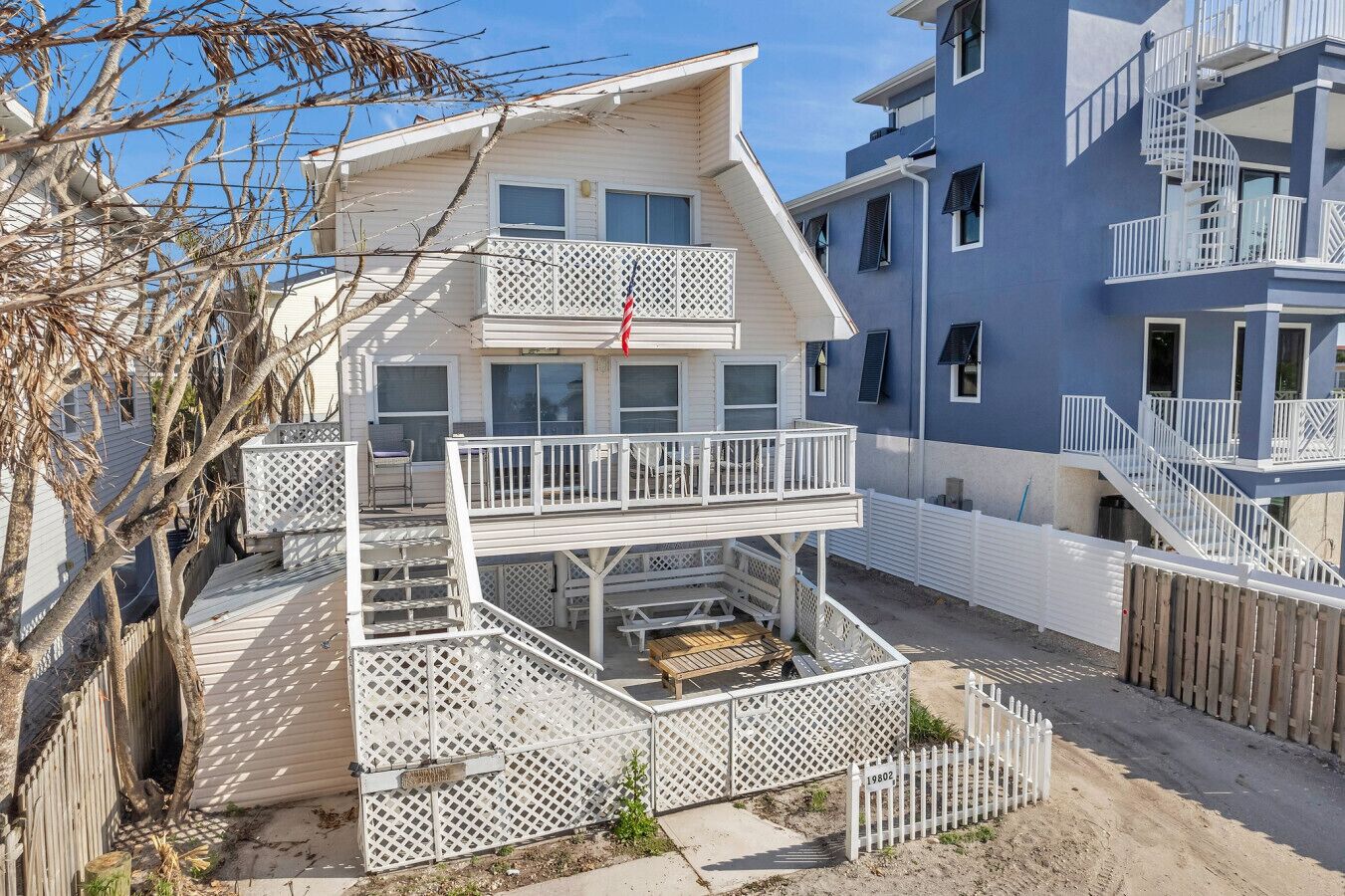 Une maison à deux étages dotée d’un balcon et d’une terrasse, entourée d’une clôture en bois et bordée d’un bâtiment bleu.