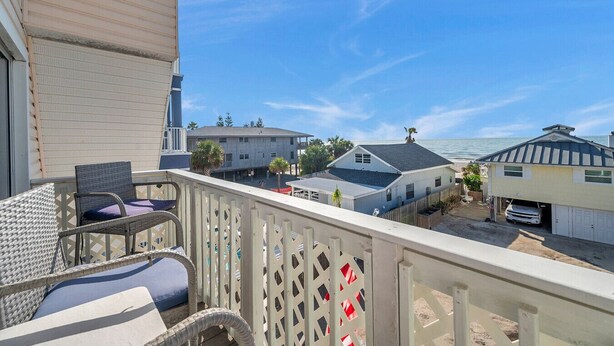 En balkon med fletmøbler, udsigt til nabohuse og en blå himmel.