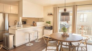 A modern kitchen with white cabinets, a stainless steel refrigerator, a microwave, and a coffee maker. There is a dining area with a round table and chairs, and a balcony with a view. - Modern Carmel Unit - Central, Big Yard & Fire Pit! (Carmel-by-the-Sea)