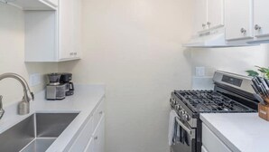 A compact kitchen with white cabinets, a stainless steel sink, a coffee maker, and a stove. - Modern Carmel Unit - Central, Big Yard & Fire Pit! (Carmel-by-the-Sea)