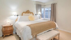 A bedroom with a large bed, bedside lamps, a wooden nightstand, a bench, and a window with curtains. - Carmel Charmer - Close to Downtown w/ Fire Pit! (Carmel-by-the-Sea)