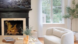 A cozy living room with a fireplace, a comfortable armchair, and a view of the outdoors through a large window. - Carmel Charmer - Close to Downtown w/ Fire Pit! (Carmel-by-the-Sea)