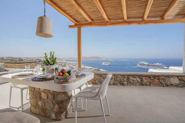 A table set with plates, glasses, and a fruit bowl, overlooking a scenic coastal view with a cruise ship in the distance.