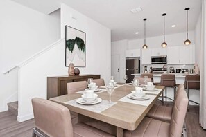 A modern dining area with a wooden table set for six, surrounded by cushioned chairs. A kitchen is visible in the background with white cabinets and stainless steel appliances. - Family 5BR Retreat Near Disney + Pool (Davenport)