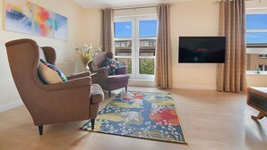A living room with a brown armchair, a sofa, a colorful rug, a flat-screen TV, and a window with curtains. - Bannermill Place Apartment | Grampian Lettings Ltd (Aberdeen)