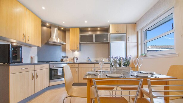 A modern kitchen with wooden cabinets, stainless steel appliances, and a dining area with a table set for four. - Bannermill Place Apartment | Grampian Lettings Ltd (Aberdeen)