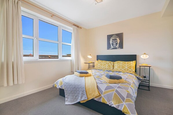 Ein Schlafzimmer mit einem Bett, zwei Nachttischlampen, einem Fenster mit Vorhängen und einer an der Wand befestigten Buddha-Statue.