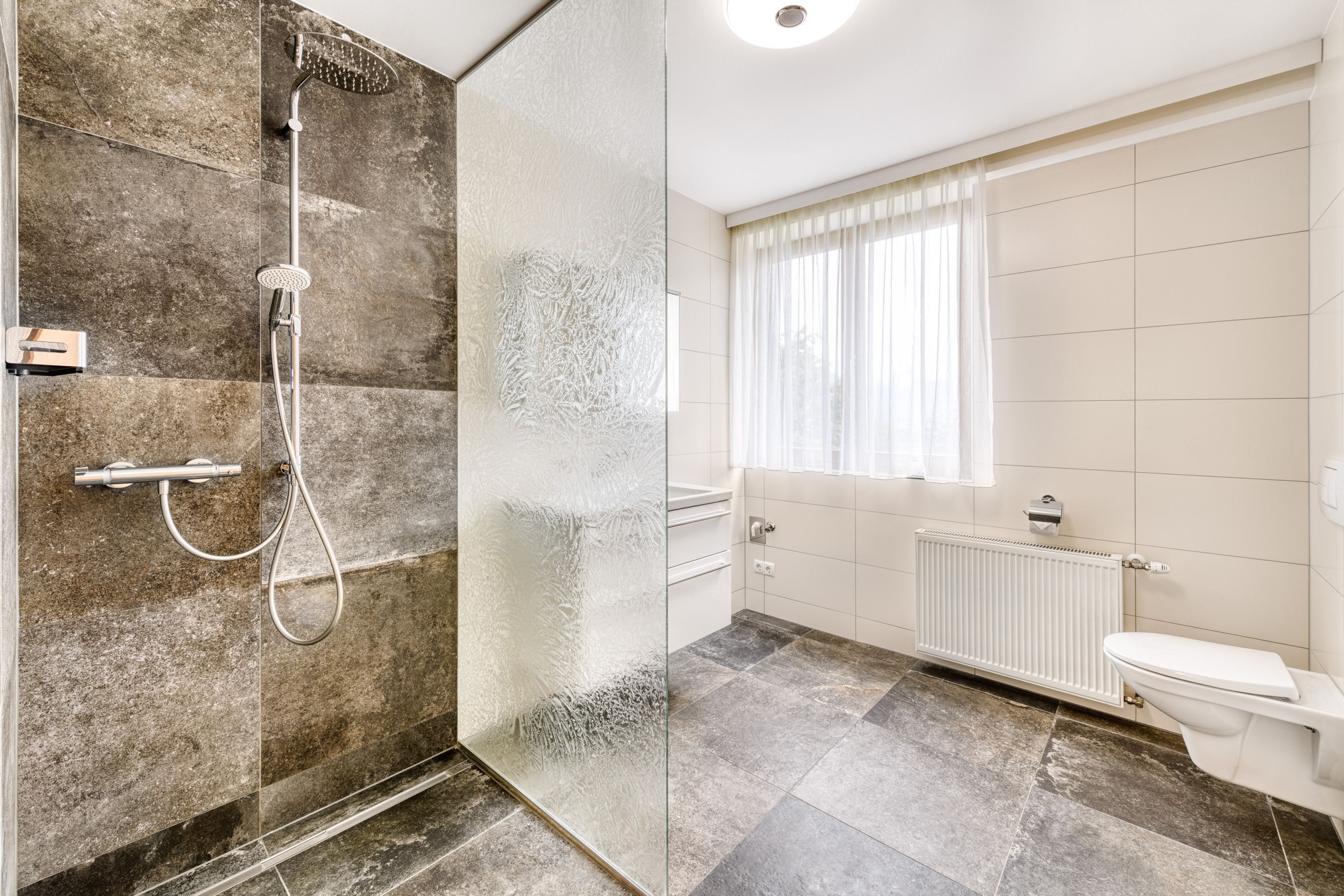 Un baño moderno con cabina de ducha de vidrio, inodoro y ventana con cortinas.