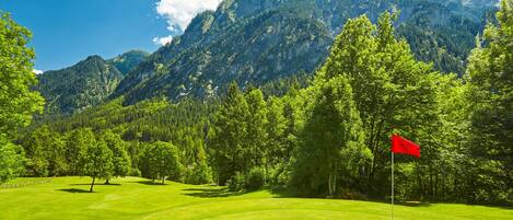 A golf course with a red flag, surrounded by lush green trees and mountains in the background.