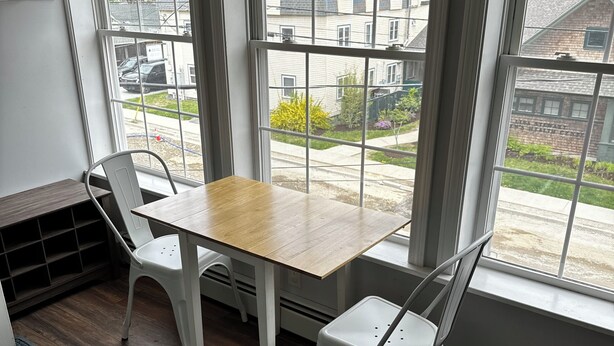 A room with a wooden table and two white chairs, large windows with blinds, a radiator, and a view of a residential area. - Charming 2-bedroom apartment with WiFi, AC in downtown Bar Harbor (Bar Harbor)