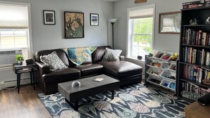 A living room with a brown leather sofa, a coffee table, and a bookshelf filled with books. - Charming 2-bedroom apartment with WiFi, AC in downtown Bar Harbor (Bar Harbor)