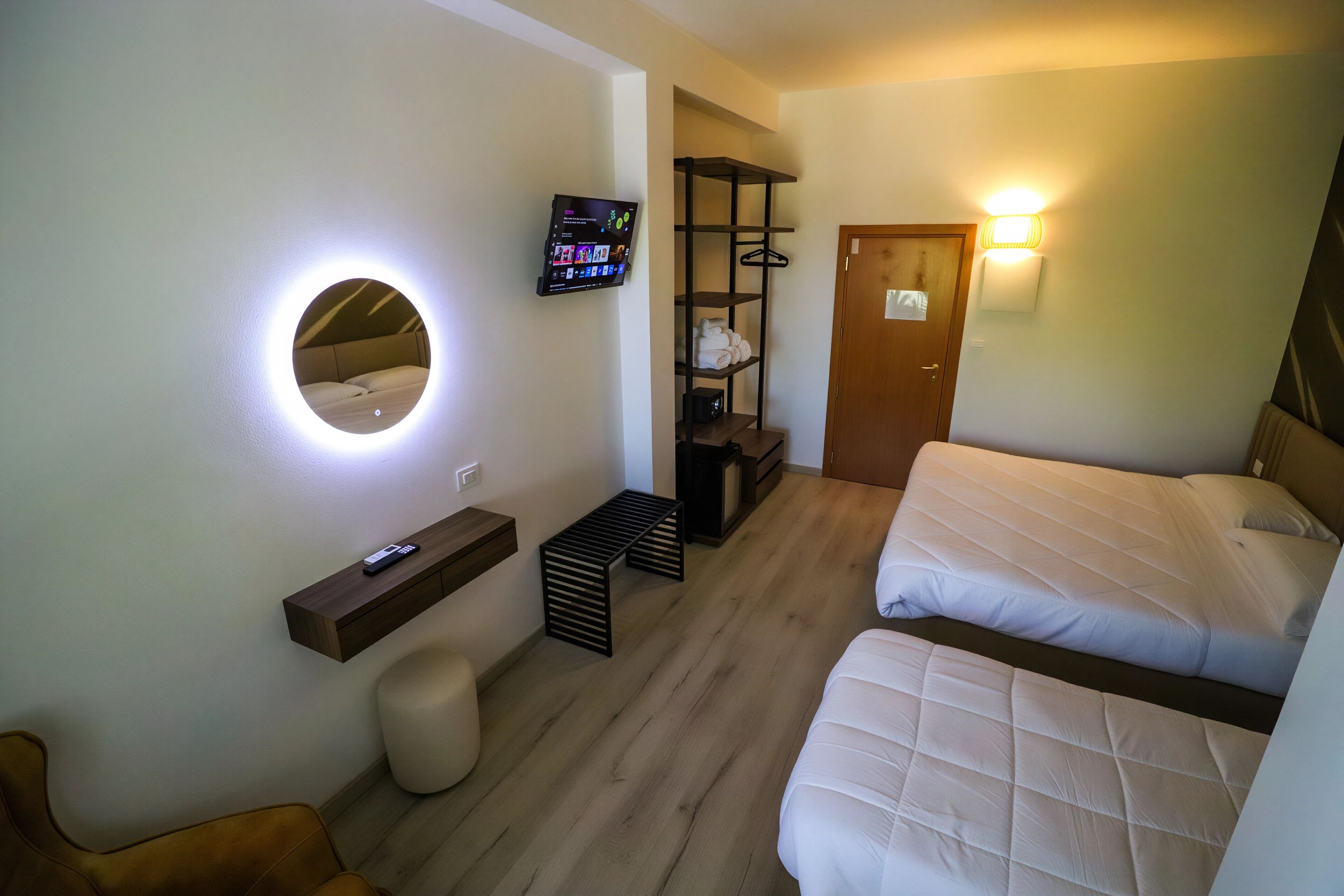 A hotel room with two beds, a TV mounted on the wall, a wooden shelf, a bench, and a door.