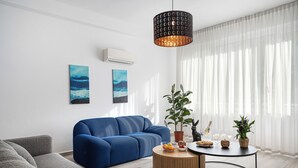 Een moderne woonkamer met een blauwe bank, een grijze bank, een ronde houten salontafel en een zwart hanglampje. Twee schilderijen hangen aan de muur, een potplant en een klein tafeltje met een fles en glazen.