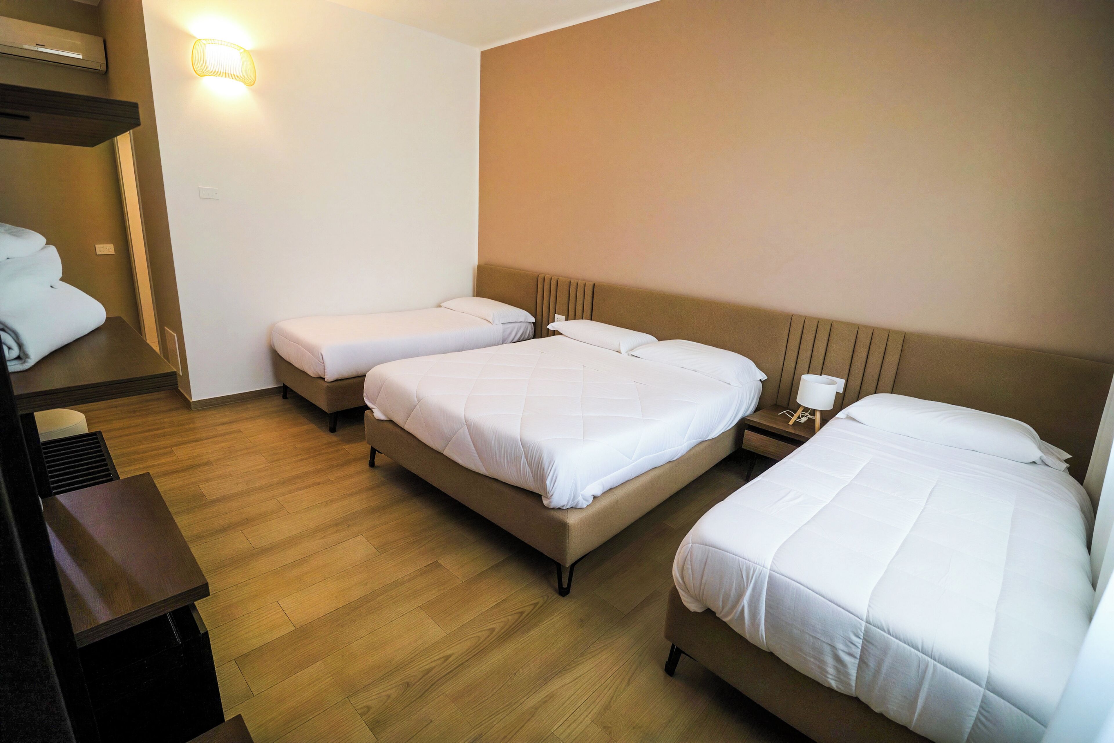 Habitación de hotel con tres camas, suelo de madera, una lámpara de pared y una mesita de noche con lámpara.