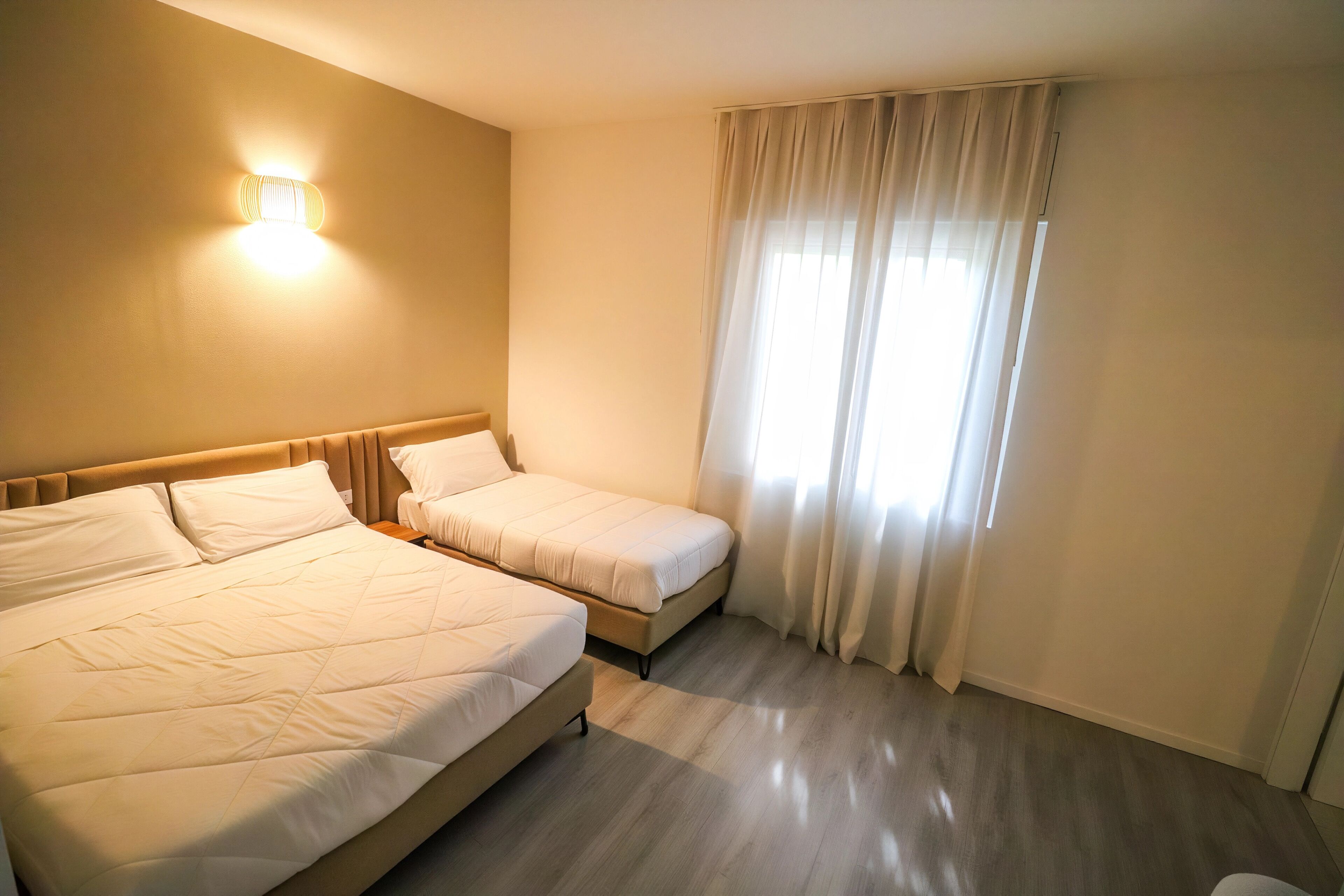 A hotel room with two beds, a nightstand, a wall-mounted light, and a window with curtains.