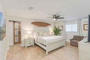 Quarto com cama, mesas de cabeceira, ventoinha no teto, janela com persianas, planta e porta com painéis de vidro.