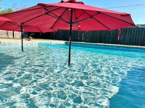 Piscina com água cristalina, cercada por uma cerca de madeira e um guarda-sol vermelho.