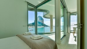 Ein modernes Hotelzimmer mit einem großen Bett, Blick auf das Meer und ein Berg, sowie ein Balkon mit einem Stuhl.