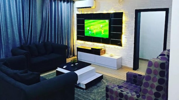 Un salon moderne avec un téléviseur à écran plat fixé sur un mur en briques blanches, un canapé noir, une console blanche et un fauteuil à motifs.