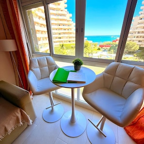 A room with two white armchairs, a small round table with a green folder and a plant, and a view of the sea through large windows.