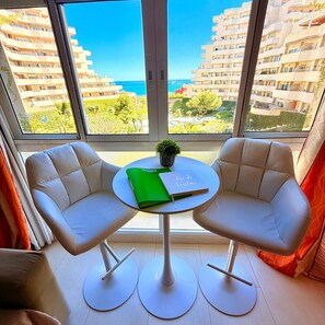 A modern room with two white armchairs, a small round table with a green folder and a notebook, and a view of a coastal cityscape through large windows.