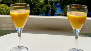To glas appelsinjuice på en balkon med udsigt til havet og bygninger.
