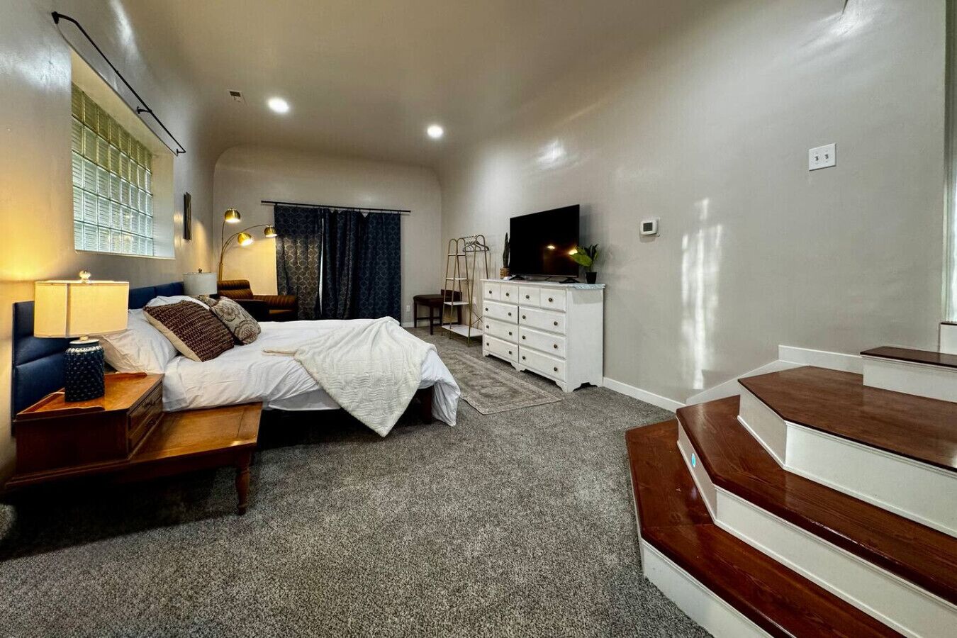 A bedroom with a bed, bedside table, lamp, dresser, TV, and a staircase.