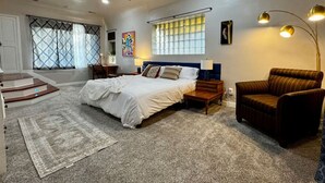 A bedroom with a large bed, a chair, a desk, and a window with blinds. - Luxurious Mid-Century-Retreat (Ogden)