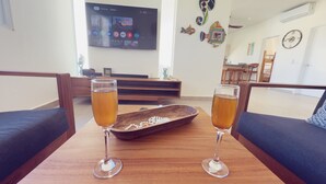Two glasses of beer on a wooden table with a decorative bowl in the center, set in a living room with a TV on the wall and a staircase in the background.