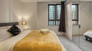 A hotel room with a bed, a nightstand, a lamp, a window with curtains, and a view of buildings outside. - Hyde Park Elegenace 2BR 2BA Balcony (London)