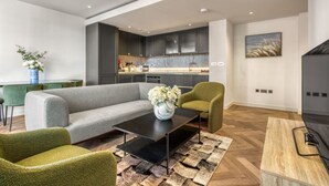 A modern living room with a grey sofa, green armchairs, a black coffee table, and a wooden side table. A kitchen with dark cabinets and a marble countertop is visible in the background. - Hyde Park Elegenace 2BR 2BA Balcony (London)