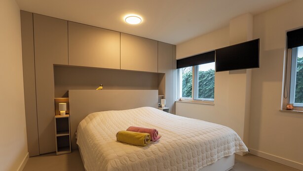 A bedroom with a bed, built-in shelving, a bedside table, a TV mounted on the wall, and a window with a view of trees.