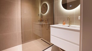 A modern bathroom with a glass-enclosed shower, a white sink with a gold faucet, and a round mirror with a light.