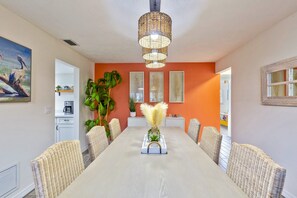 A dining area with a long table, wicker chairs, a hanging light fixture, and a painting on the wall.