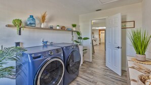 Un área de lavandería con lavadora y secadora, un estante con objetos decorativos y una puerta que da a otra habitación.