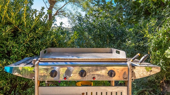 A barbecue grill with a flat top, four burners, and a side burner, set against a backdrop of dense foliage.