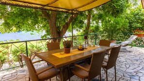 A covered outdoor seating area with a table and chairs, surrounded by greenery and overlooking a body of water.