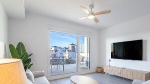 Un salon moderne doté d’une grande porte-fenêtre coulissante donnant sur un balcon, d’un ventilateur de plafond, d’un téléviseur à écran plat fixé au mur et d’un meuble en bois au design minimaliste.