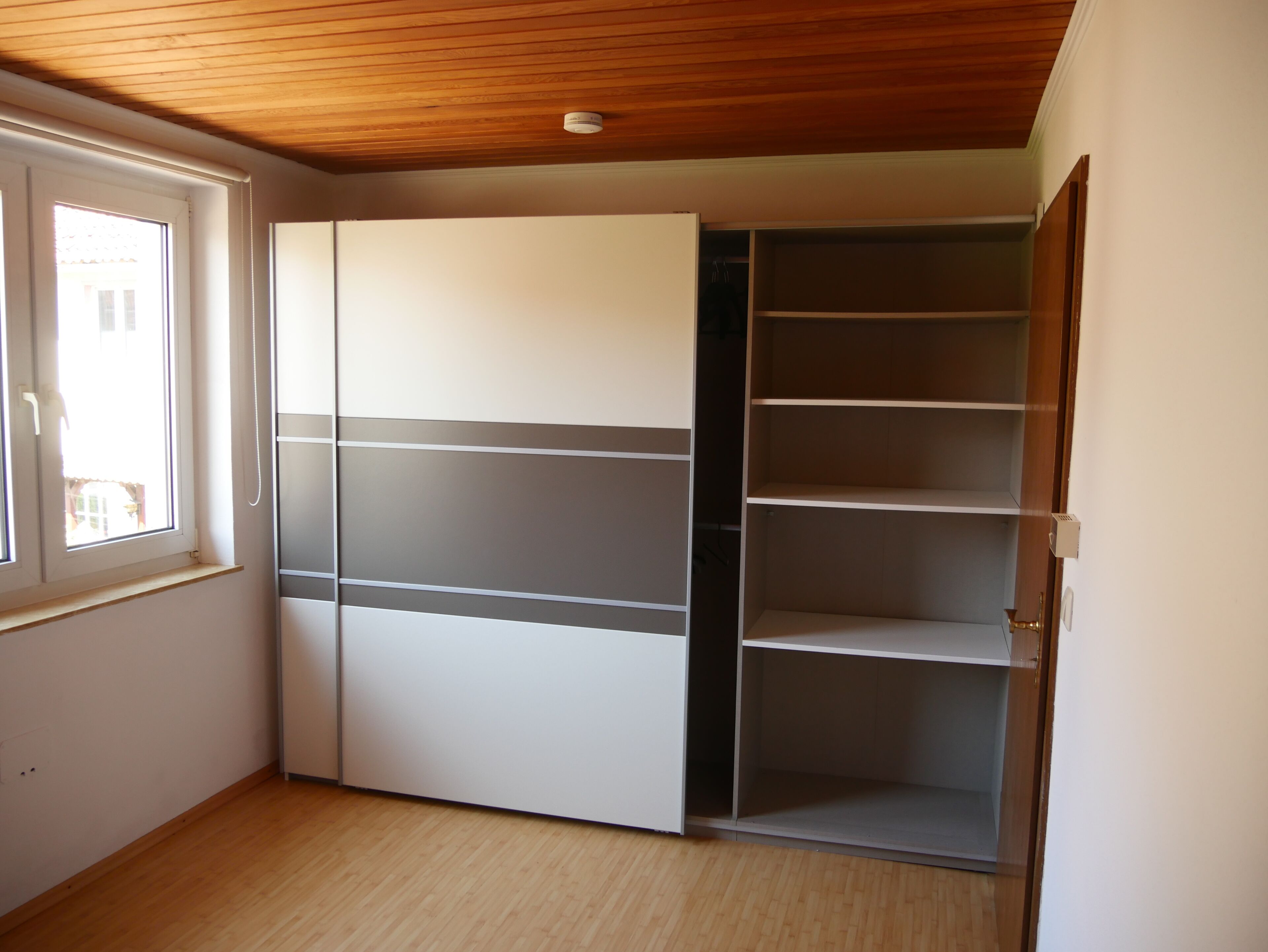 A room with a wooden ceiling, a white wardrobe with grey horizontal stripes, and a window with a view of buildings outside.